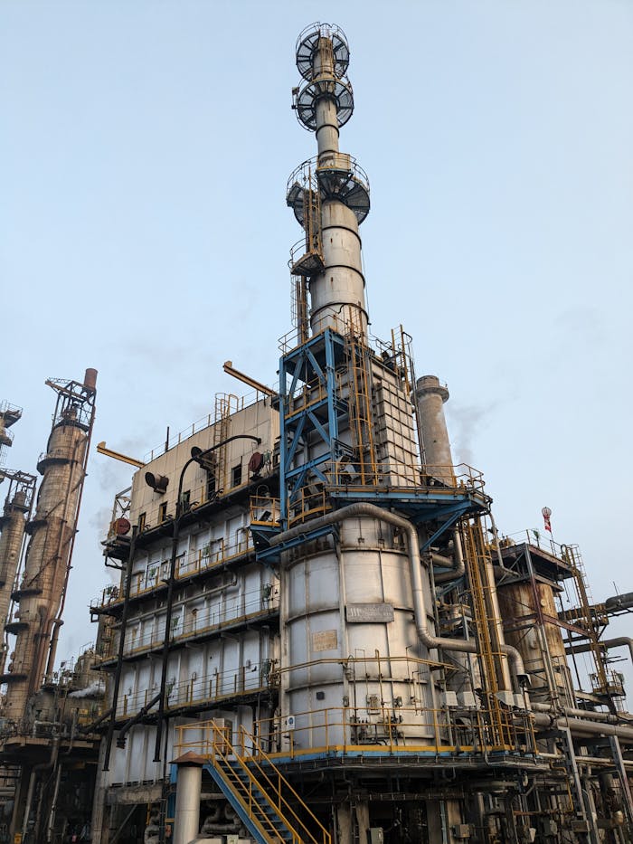 Detailed view of a large industrial oil refinery tower with intricate piping and structures against a clear blue sky.